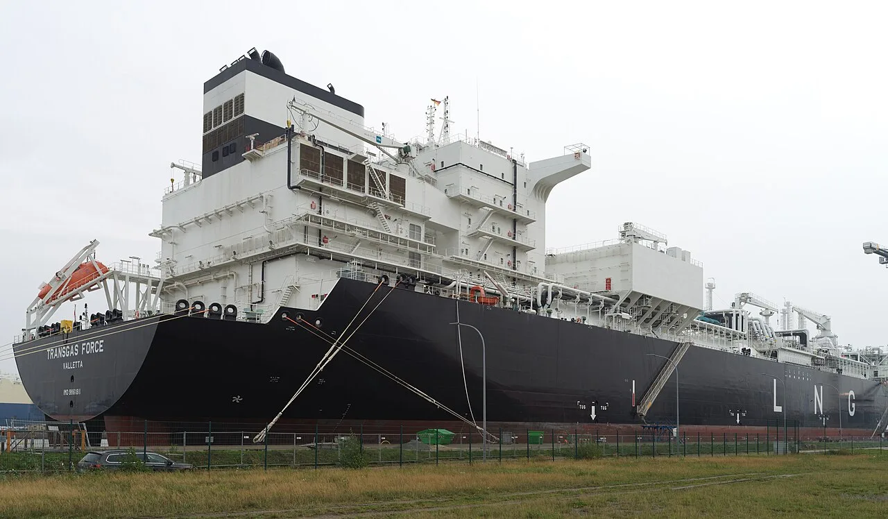 Velký tanker na LNG s černým trupem kotvící u nábřeží, s bílou nástavbou, potrubím a nápisem LNG na boku.