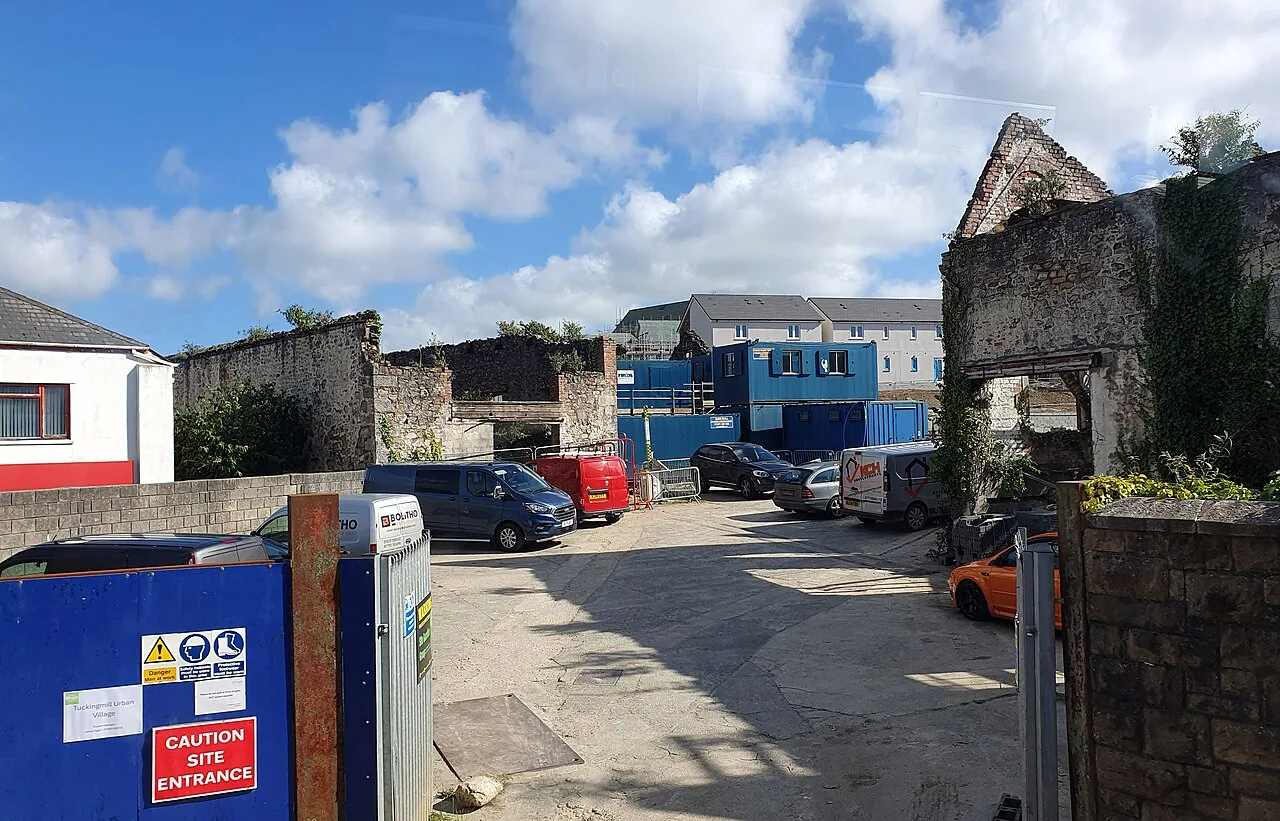 A construction site with parked vans, blue site cabins and ruined stone walls, with new houses rising behind under a blue sky.
