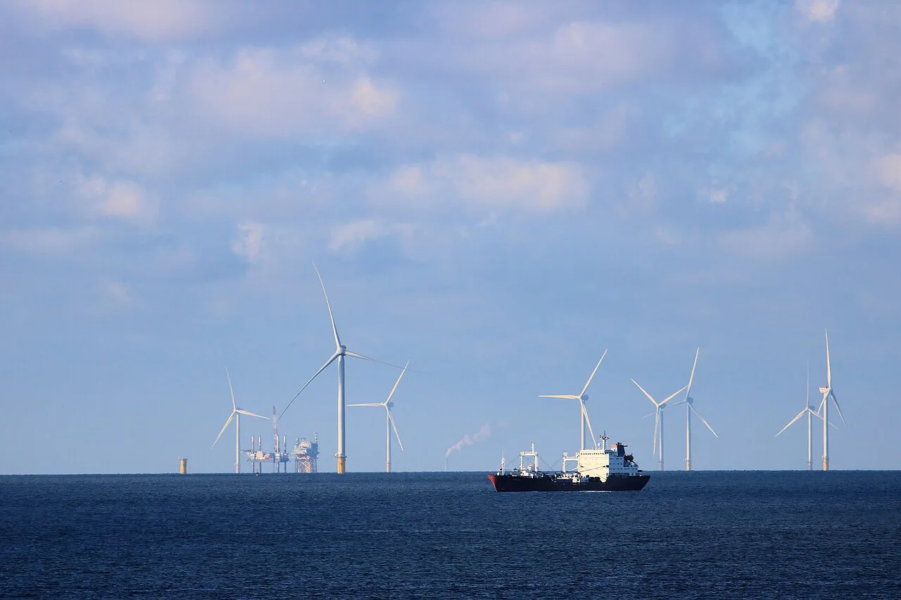 A dark-hulled tanker on calm blue sea with offshore wind turbines and a platform on the horizon under a cloudy sky.
