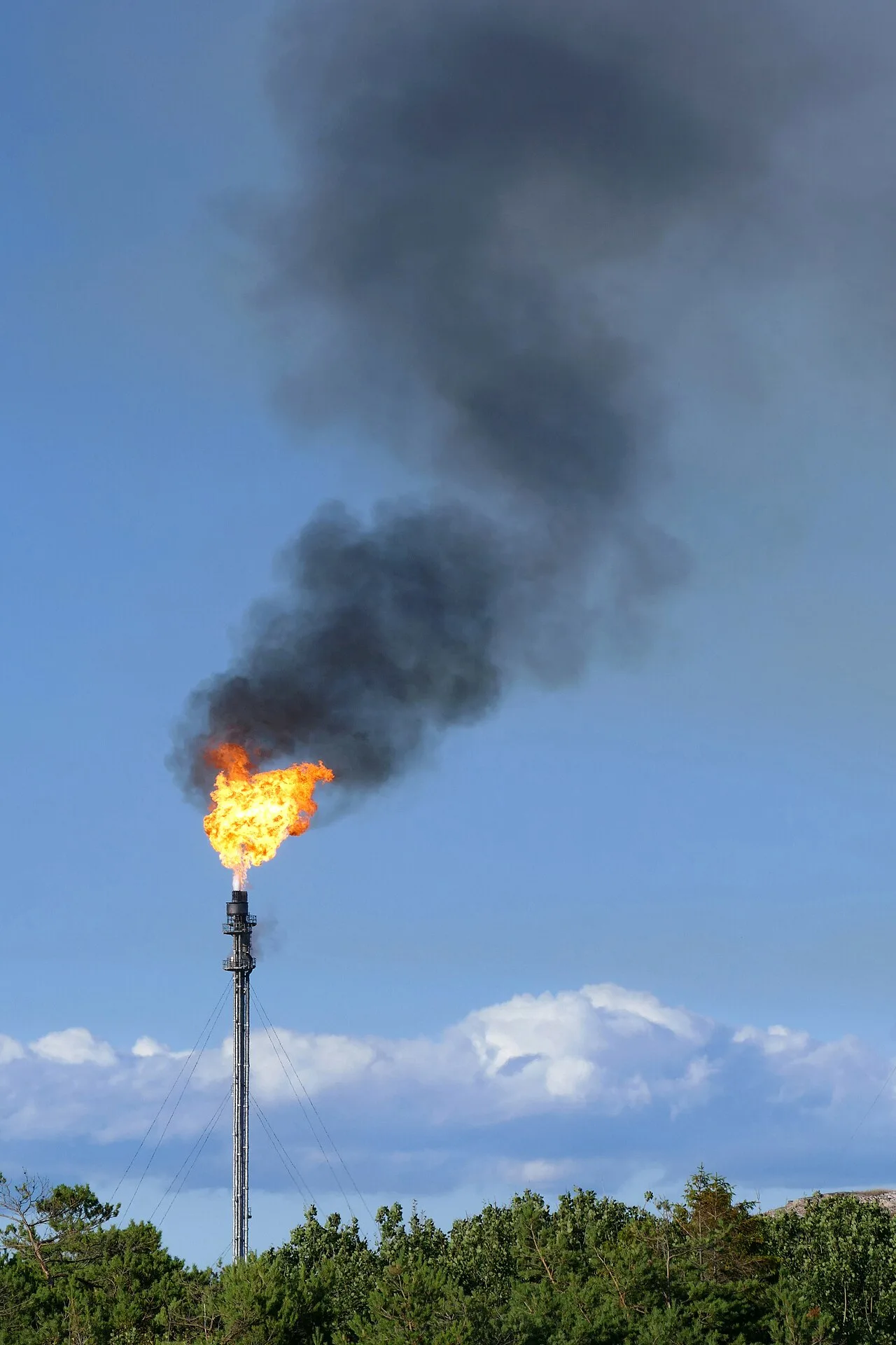 A flare stack burning a bright flame with black smoke drifting across a blue sky above a treeline.