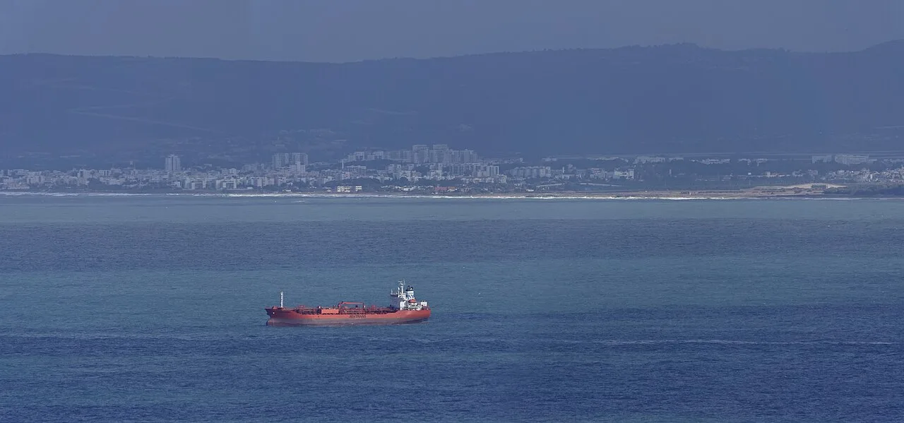 Ein roter Öl- und Chemikalientanker liegt in blauen Küstengewässern vor Anker, dahinter eine dunstige Stadtsilhouette und Hügel.