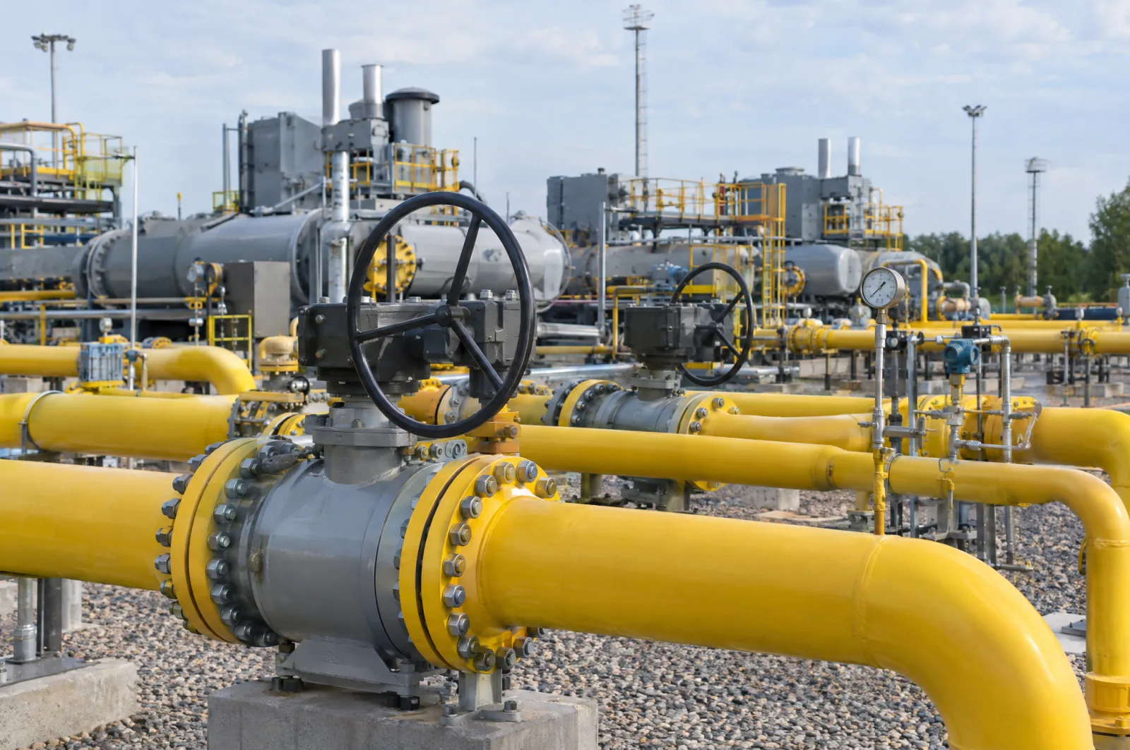 Grote grijze kleppen met zwarte handwielen op felgele gasleidingen bij een compressorstation in de open lucht op een grindterrein bij daglicht.