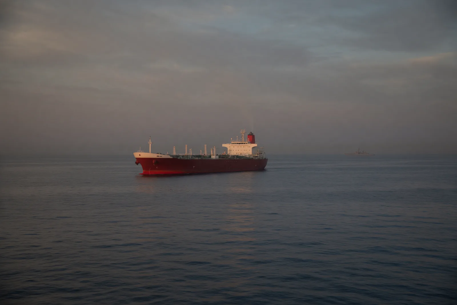 A red-hulled oil tanker alone on calm grey water at dusk, a faint warship silhouette on the hazy horizon.