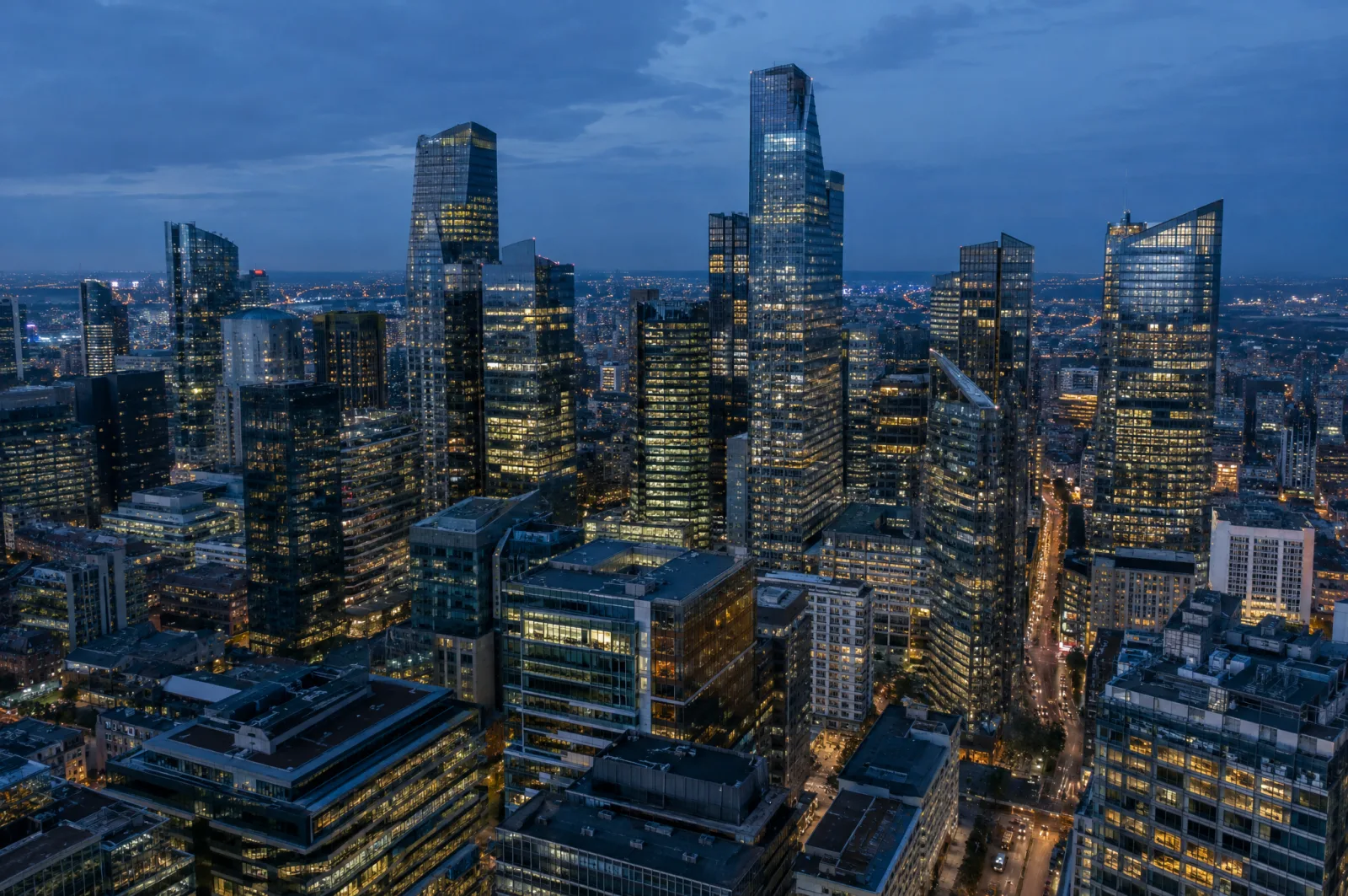 An aerial view of a dense district of glass skyscrapers at blue hour, office windows lit warm against a deep blue sky.