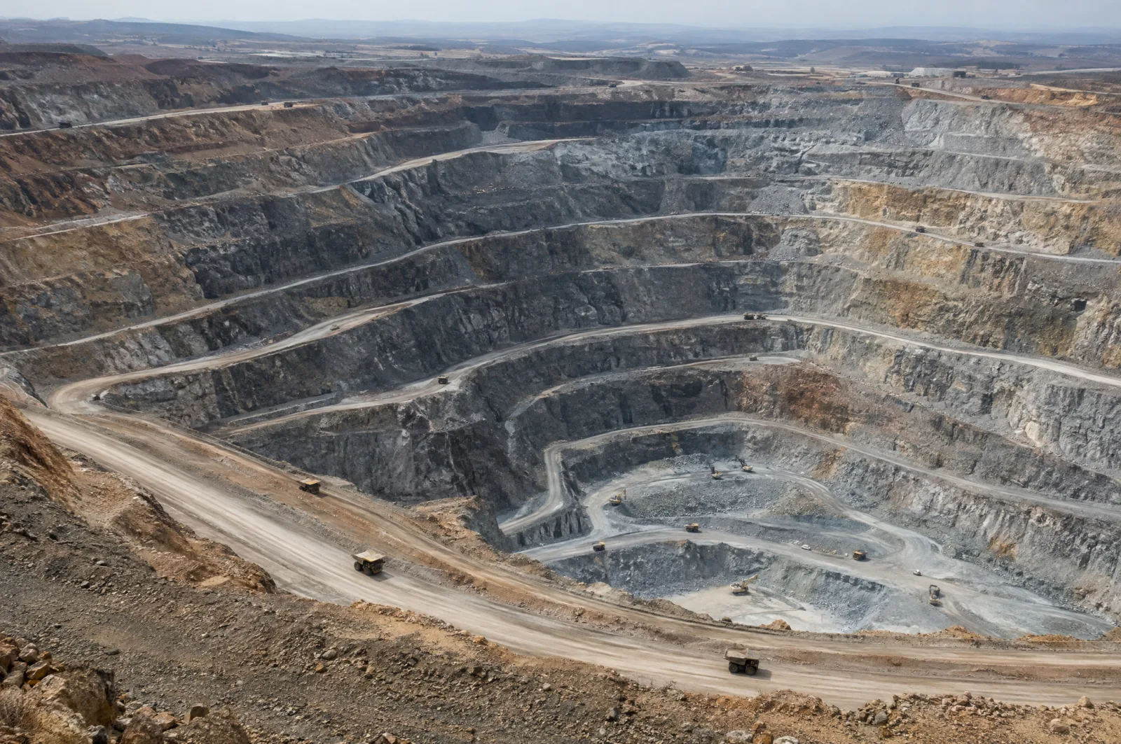 A huge terraced open-pit mine seen from above, with haul roads spiralling down its grey and ochre walls and small trucks on the ramps.