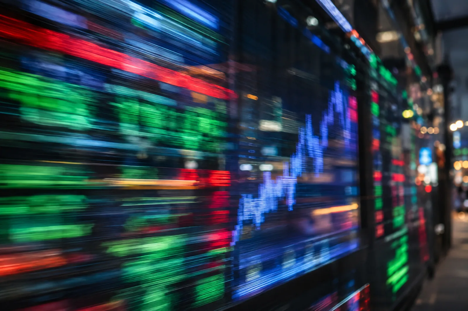 Motion-blurred red, green and blue lights of an electronic market display wall at night, with a glowing line chart in the middle.