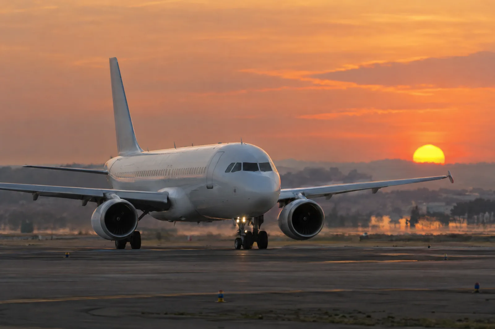 A plain white passenger jet faces the camera on a runway as a large orange sun sets behind it.